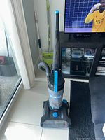 Bissell PowerForce upright vacuum cleaner positioned upright on tiled floor next to black carpet and couch, showing front and side with hose and handle visible.