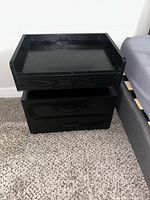 Front view of the black wooden nightstand showing the top flat surface with raised edge and power outlet with switches, two spacious drawers with black handles, placed on a carpeted floor beside a bed.