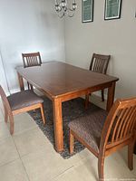 Photo showing the rectangular wooden dining table with gloss finish and four matching wooden chairs with fabric cushions around it, placed on a dark rug on tiled floor.