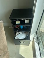Black wooden side table with one drawer open and an open shelf, placed on tiled floor next to glass door and couch.