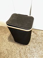 Closed black fabric laundry basket with lid placed upright on carpeted floor.
