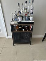 Full front view of the medium liquor cabinet showing wood and metal construction, a top surface with bottles (not for sale), and bottom enclosed glass door storage.