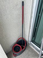 Full mop and bucket set resting against a textured wall near a sliding door. The bucket is gray with red handles and a built-in wringer, featuring wheels for easy movement.