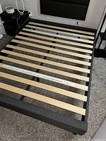 Full view of queen bed frame showing natural wood slats on black metal frame against carpeted floor, alongside nightstand and wall outlet.