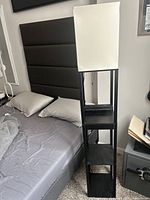 Full view of the floor lamp next to bed showing square fabric shade, black frame, and shelves
