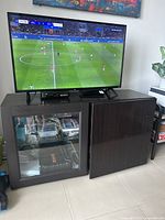 Full front view of dark wooden TV stand with flat screen TV on top. Left door is glass showing shelves with items inside. Right solid door partially closed.