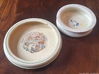 Two round ceramic children’s bowls on wooden table, larger bowl shows illustration and smaller bowl shows wear and crack on bottom