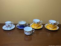 Five small antique cups and four matching saucers arranged in one row on a flat surface against a plain wall background