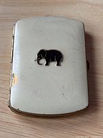 Front of the brass cigarette case showing white enamel coating and black elephant design with signs of wear and enamel chipping