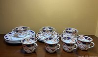 Seven cups with matching saucers and decorated plates arranged in rows against a beige wall background.