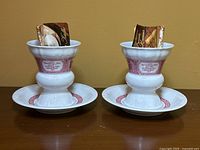 Two white porcelain coffee and dessert cups with pink decorative bands sitting on matching saucers. Each cup holds an instruction inset card inside.