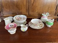 View of seven floral-themed China pieces arranged on a wooden cabinet, showing their varied shapes and detailed floral patterns.