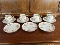 Four limoges porcelain demitasse cups on four matching saucers with an additional three matching saucers laid out on a wood surface. Each cup is white with pink and gold floral pattern and gold trim.