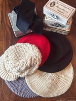 Six knit hats in cream, gray, red, black, beige (five flat style and one chunky cable knit) with a black neck tie and three vintage advertising boxes placed on wooden surface