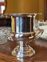 Front and side view of the polished silver plated champagne bucket showing the shape, twin handles, and pedestal base on a wooden surface.