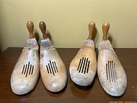 Front view of two pairs of wooden shoe trees on wooden table against beige wall.
