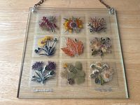 Full view of the glass square hanging art showing nine pressed flower designs arranged in three rows, with hanging chain attached at two corners. Signed by the artist on lower left and right corners.