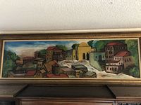 Full view of the horizontal abstract cityscape painting in an ornate wooden frame hung on the wall, showing colorful buildings, greenery and sky.