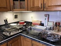 Wide view of kitchen countertop displaying assorted kitchenware items including cutlery, steamer, casserole dish, baking dishes, and kitchen utensils showing arrangement and variety.