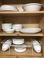 Shelves inside a cabinet displaying white Corelle Vitrelle glass dinnerware including plates, bowls, and mugs