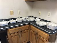 Set of multiple CorningWare Blue Cornflower casserole dishes arranged on countertop including rectangular, square, and round shapes with lids and some small bowls without lids