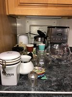Photo shows Gevalia branded coffee maker with glass carafe wrapped in plastic and several white ceramic canisters, a white teapot, small cups and various small accessories on dark kitchen countertop under wooden cabinets.