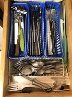 Top view of kitchen utensils organized in three blue plastic trays containing forks, knives, and other cutlery with additional spoons and utensils in front.