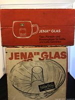 Two original boxes stacked, top box for six tea glasses, red with Schott Mainz Jenaer Glas branding, bottom box for glass bundt pan with illustration and text 'Jenaer Glas'