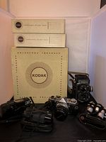 Overview of three Kodak Carousel slide tray boxes at back, Rolleiflex camera at right, Olympus OM-1 and Trip Zoom 230 cameras in front, projector lens and cases at left
