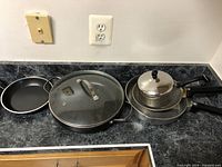 Photo showing two metal frying pans, a large metal pan with glass lid, and small metal saucepan with lid, all with black plastic handles.