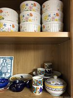 Shelves with various ceramic tableware including ramekins stacked, small cups, a blue fish-shaped dish, and a lidded bowl with floral patterns.