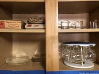 Two shelves inside a wood cabinet showing several clear glass plates stacked and several glass cups on a rack.
