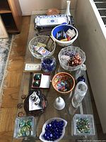 Photo shows a glass table with various decorative bowls, glass marbles, a blue ceramic bowl, small decorative items, a tall white vase, and a rectangular white and blue Italian ceramic platter.