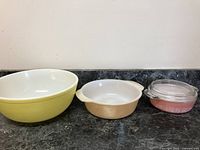 All three vintage bowls and casseroles grouped together on countertop. Yellow Pyrex bowl, peach Fire King casserole, pink Pyrex Gooseberry casserole with lid.
