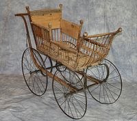 Front-side view of antique baby carriage showing wooden frame, metal spoked wheels, curved handles and decorative sides.