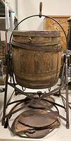 Full view of wooden barrel butter churn on metal stand with handle and visible metal band separation and damaged base pieces placed below.