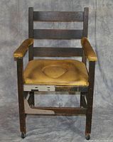 Front view of yellow commode chair with oak frame, wooden slat back, armrests with padded cover, and padded seat with removable center insert showing wear and stains.