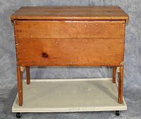 Front view of wooden dough box on four legs with removable lid, showing aged wood and chipping varnish.