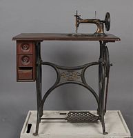Full view of antique Raymond sewing machine on wooden table with black cast iron treadle base and three drawers on left side. Visible pedal and handwheel.