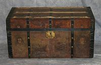 Closed wooden and leather travelling trunk showing black metal reinforcements, brass lock, and overall surface wear.