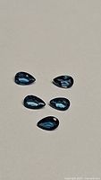 Five pear cut deep blue natural London blue topaz gemstones shown from above on plain surface.
