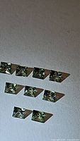 Ten princess cut natural pastel mossy green sapphires arranged on grey textured surface, showing clarity and uniform square shape sides.