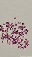 Close-up photo showing numerous small round pink sapphires with diamond cut facets, about 1mm size each, arranged loosely on a flat surface.