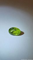 Photo of oval cut apple green peridot resting on white surface under natural light showing color and faceting details.