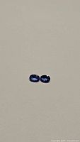 Pair of deep blue-purple oval cut tanzanite gemstones on neutral background.