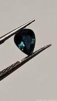 Photo of natural blue teal sapphire pear cut gemstone held by tweezers showing two angles displaying color and clarity.