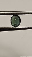 Close-up photo of oval cut blue-green sapphire gemstone held by tweezers showing color, shape and clarity.