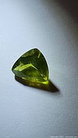 Close-up photo of natural apple green peridot, trillion cut gemstone with visible facets and vibrant green color, photographed on white background.