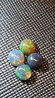 Four calibrated 4mm round Ethiopian black opals with vibrant rainbow flash laid out on a textured black surface, showing varying colors and light play in different lighting angles.