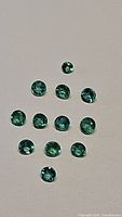 Thirteen small, round cut emerald gemstones laid out on a neutral background showing size and color.
